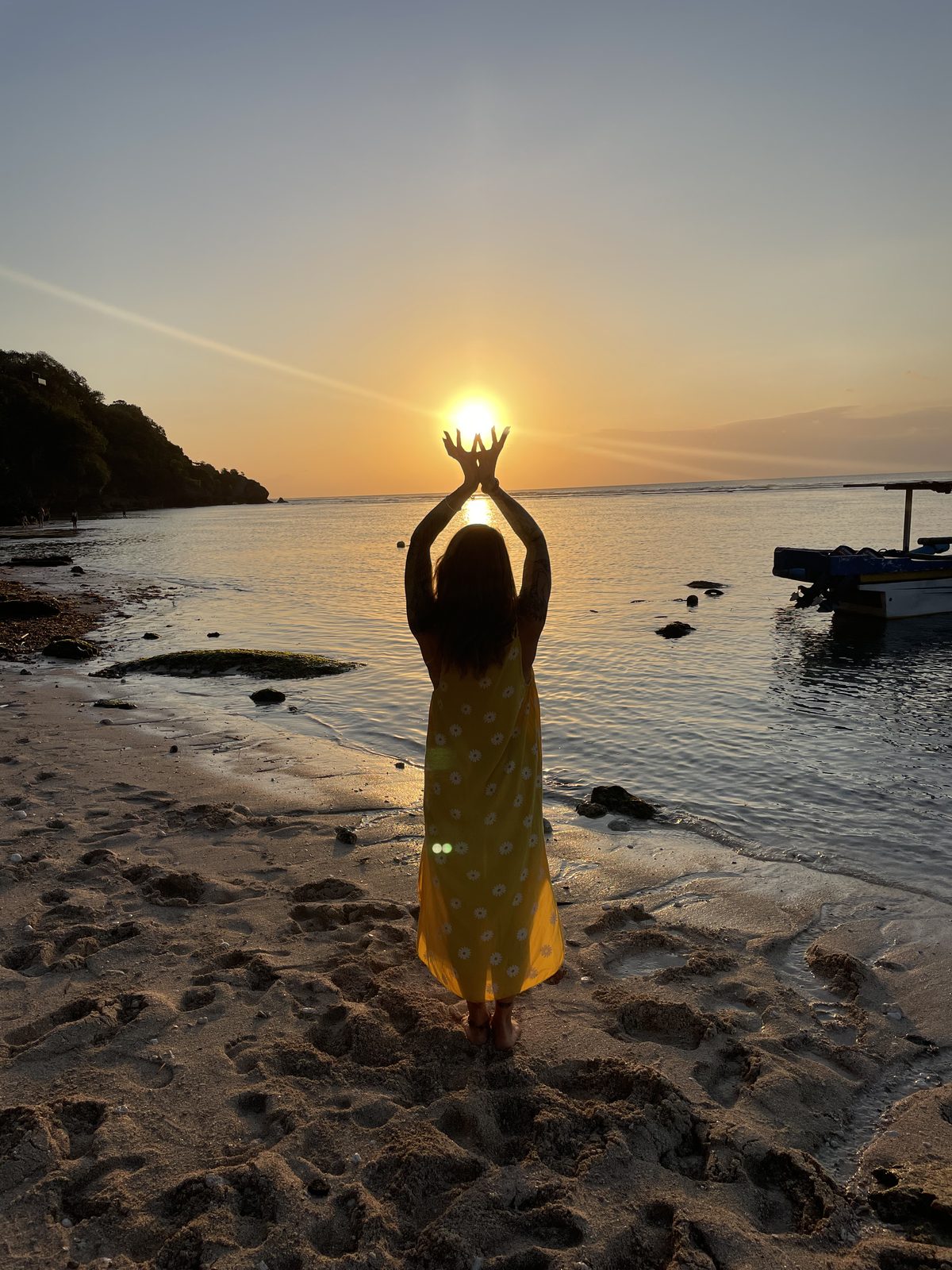 Cassandre debout sur une plage au coucher du soleil, bras levés, encadrant le soleil entre ses mains — photo prise à Bali